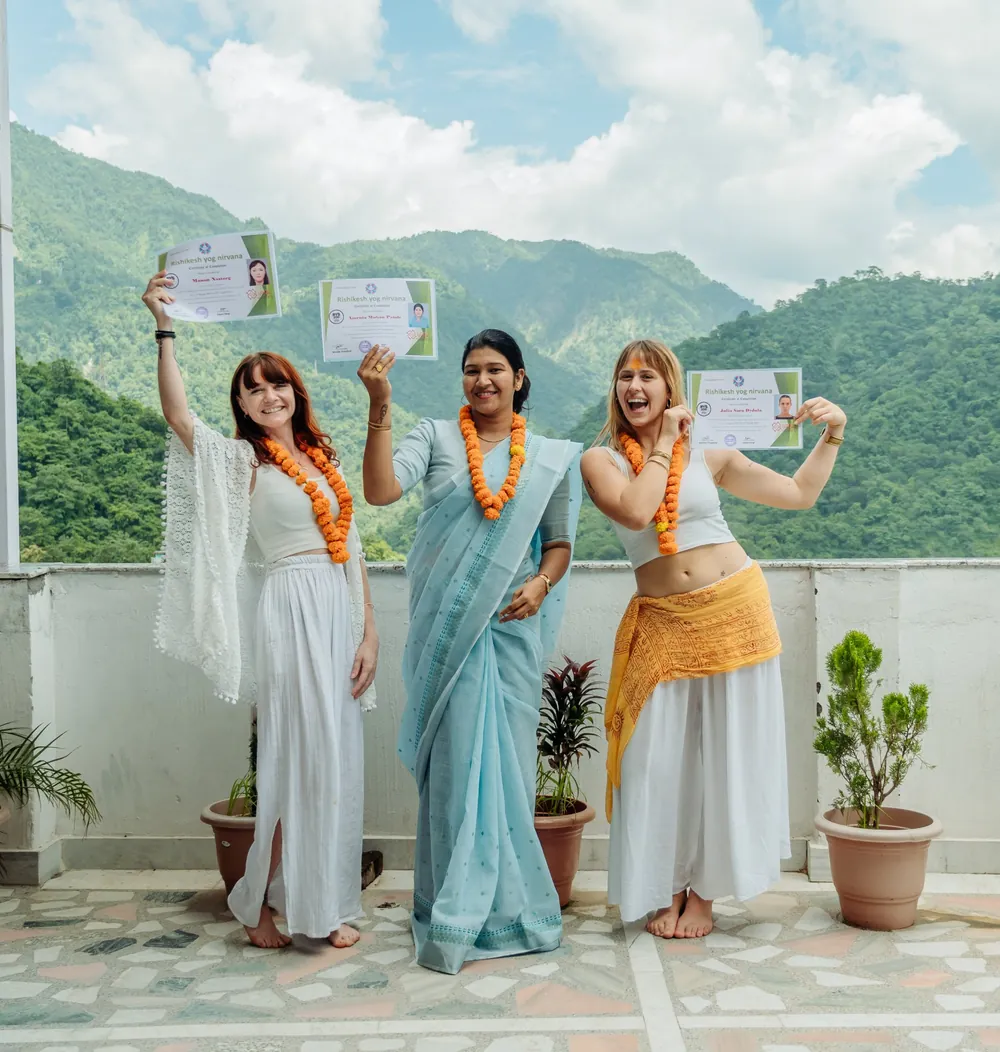 Three graduates holding up their yoga teacher training certificates on the school rooftop, with the Rishikesh hills behind them