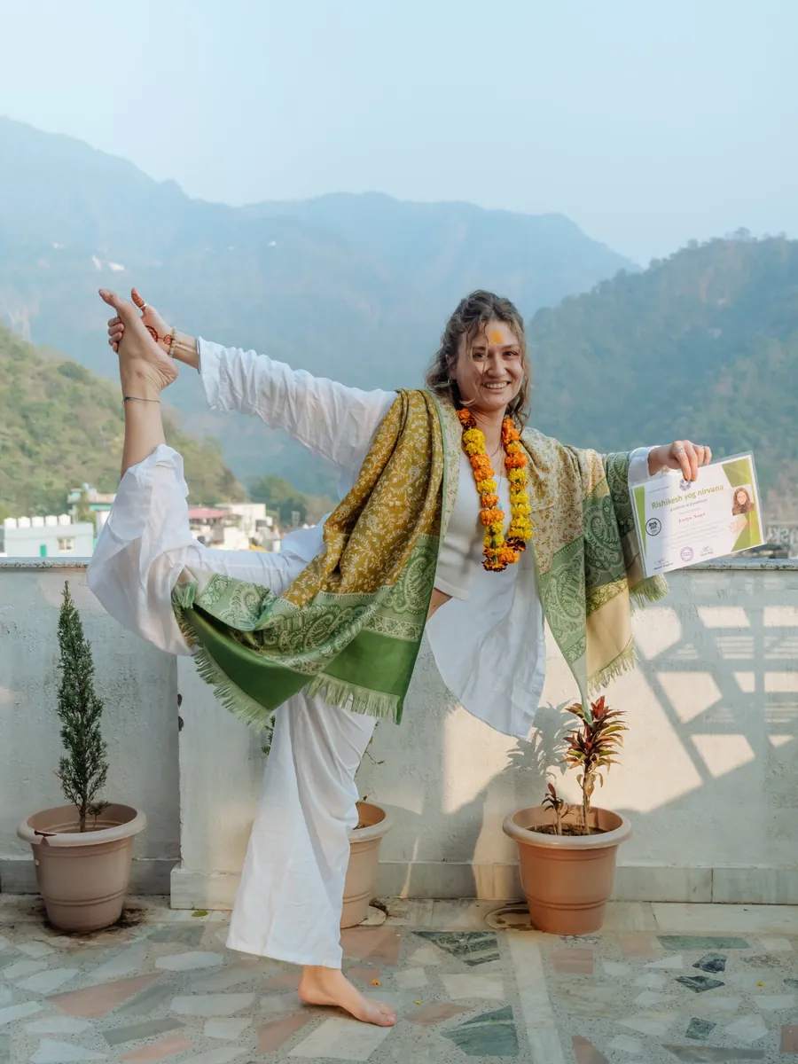 A graduate holding her yoga teacher training certificate while balancing in a standing pose on the school rooftop in Rishikesh